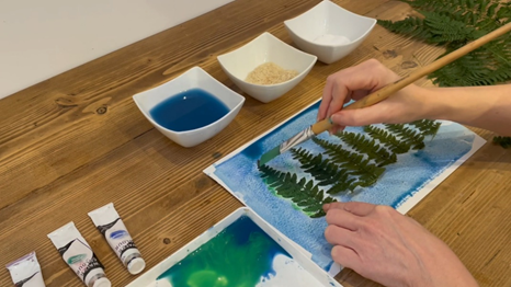 A hand with a paintbrush, painting a watercolour wash on top of leaves
