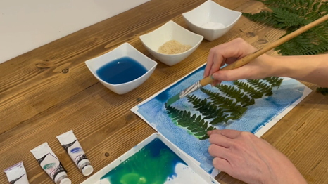 A hand with a paintbrush, painting a watercolour wash on top of leaves 